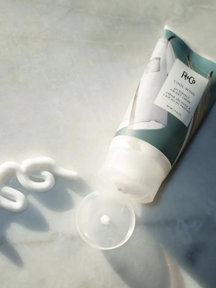 Tube of R+Co 'COOL WIND pH Perfect Air Dry Creme' next to a product swatch on a a marble background, 147ml | Twentyseven Toronto