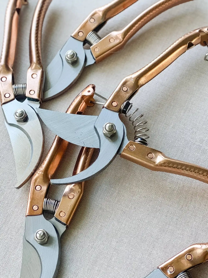 Multiple Copper Handle Garden Secateurs from Laura Hooper Design House are scattered across a neutral fabric, with one pair open to reveal the spring-action mechanism and curved cutting edge | Twentyseven Toronto
