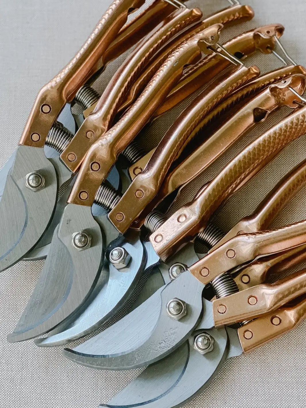 A collection of Copper Handle Garden Secateurs from Laura Hooper Design House are lined up closely together, showcasing their metallic rose-gold finish and sharp steel bypass blades | Twentyseven Toronto