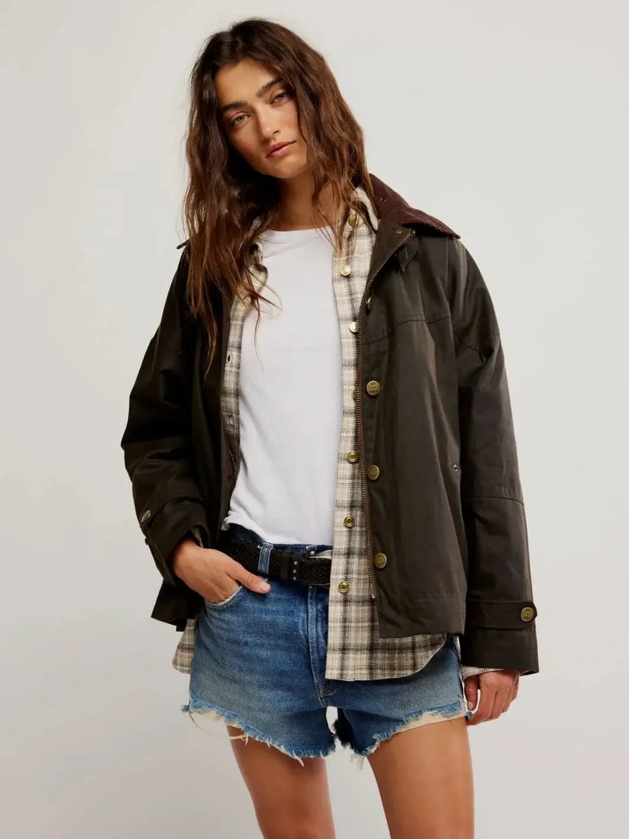 A studio image captures a model wearing the Free People Cori Waxed Jacket in Olive layered over a plaid shirt and distressed denim shorts | Twentyseven Toronto