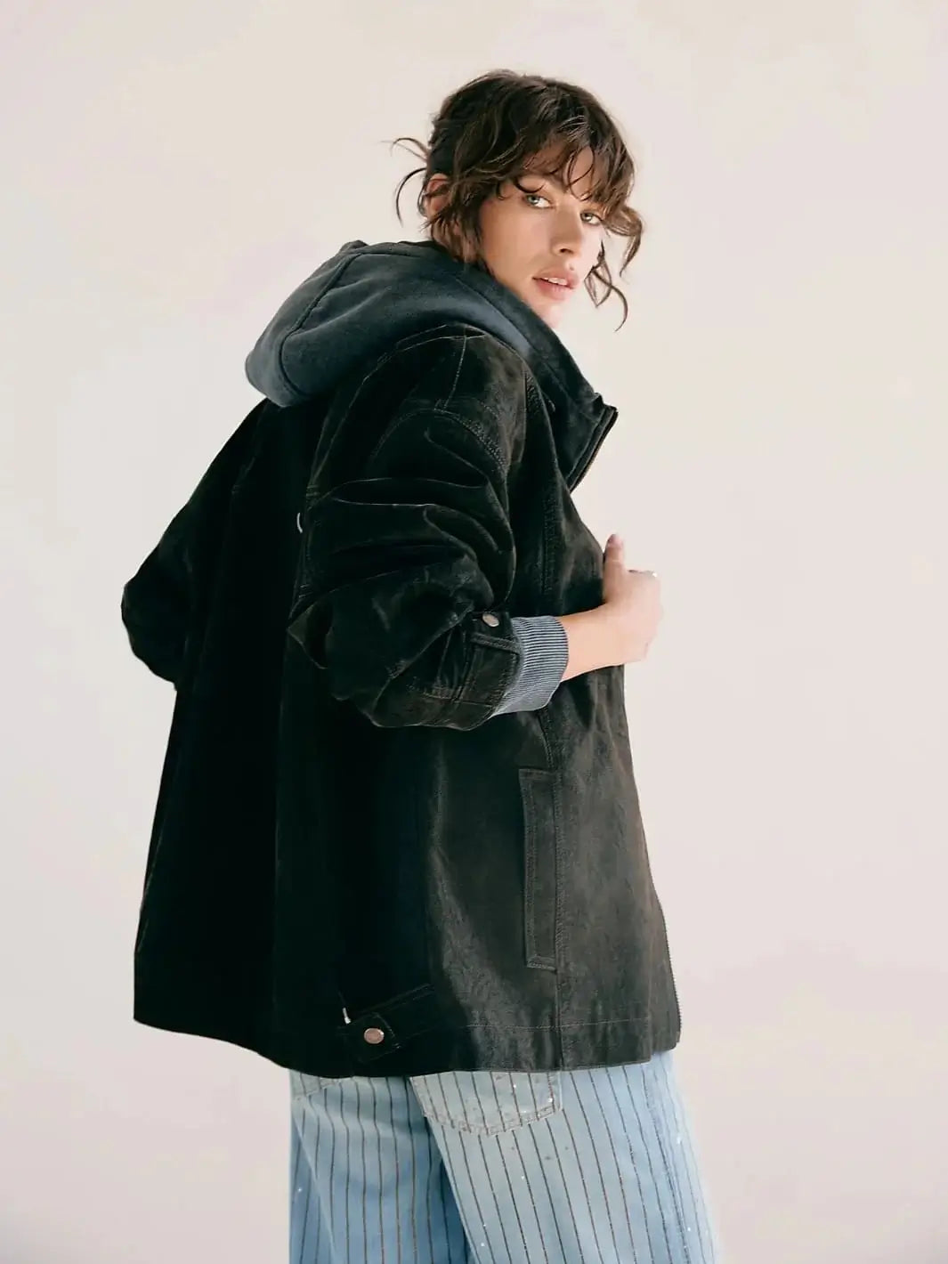 A side profile view displays a model wearing the Free People Cori Waxed Jacket in Olive styled with a grey hooded sweater underneath and striped pants | Twentyseven Toronto