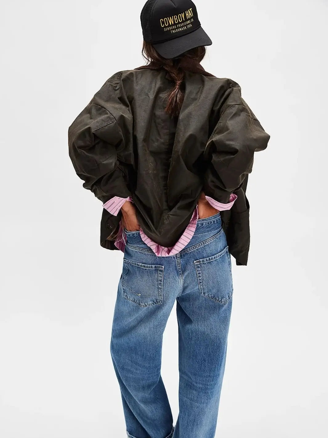 A rear view captures a model wearing the slouchy silhouette of the Free People Cori Waxed Jacket in Olive paired with a pink striped shirt, blue jeans, and a black cap | Twentyseven Toronto