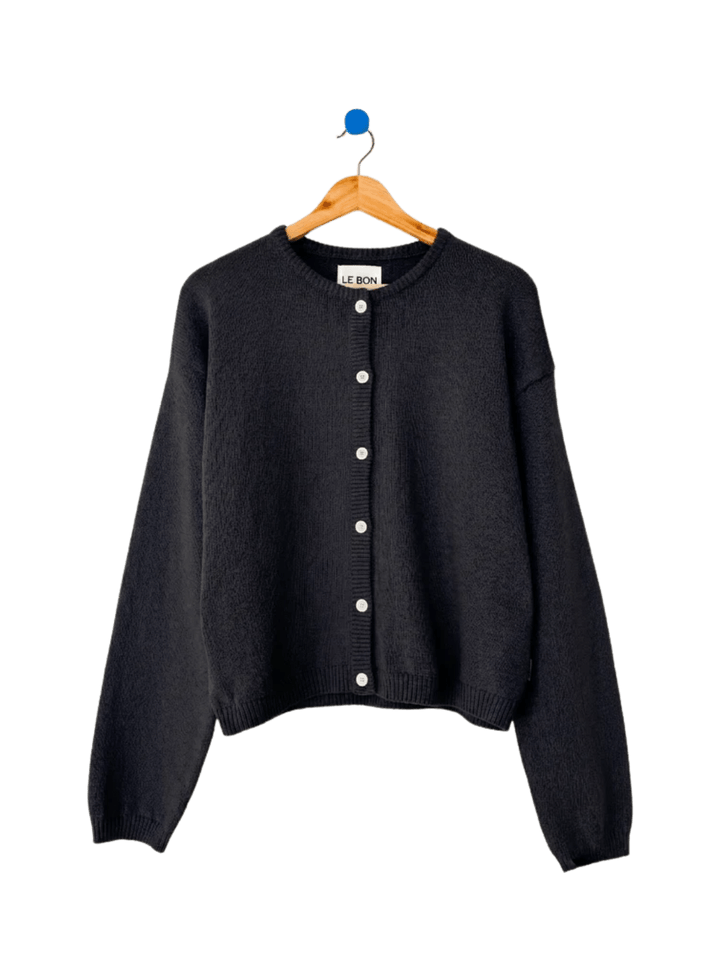 A flatlay image displays the classic button-down design and crew neckline of the Le Bon Shoppe Cotton Leila Cardigan - Black hanging on a wooden hanger | Twentyseven Toronto