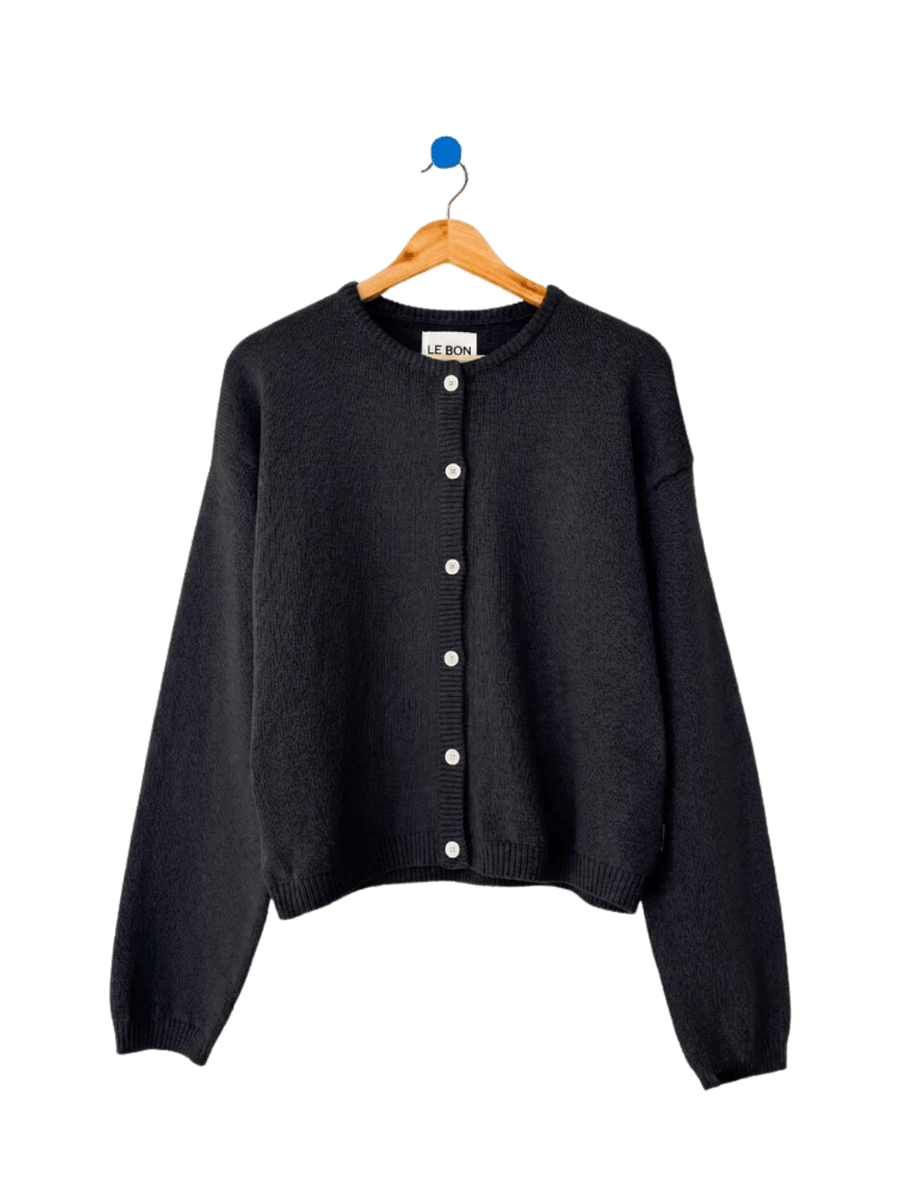 A flatlay image displays the classic button-down design and crew neckline of the Le Bon Shoppe Cotton Leila Cardigan - Black hanging on a wooden hanger | Twentyseven Toronto