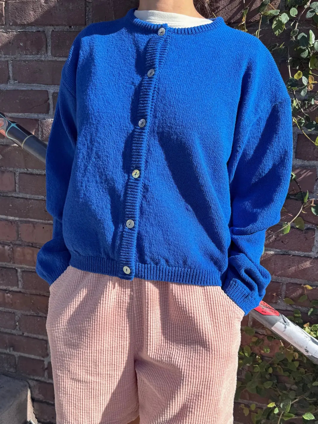 Person wearing a blue 'Cotton Leila Cardigan' in 'Cobalt' from Le Bon Shoppe against plants and a brick wall | Twentyseven Toronto