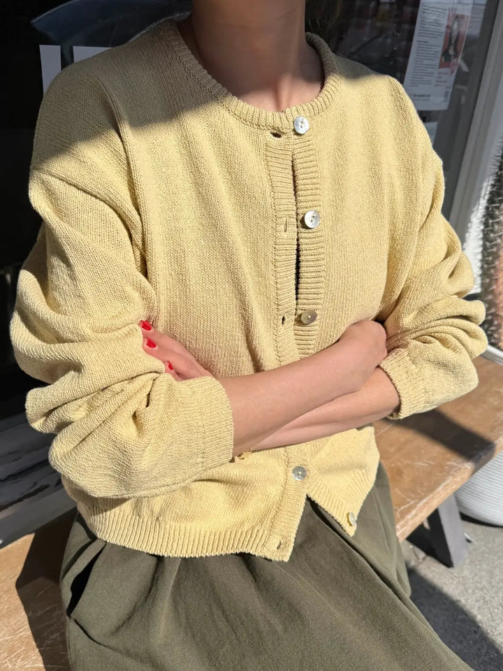 Person wearing a light yellow 'Cotton Leila Cardigan' in 'Custard' from Le Bon Shoppe with button details, sitting outdoors | Twentyseven Toronto