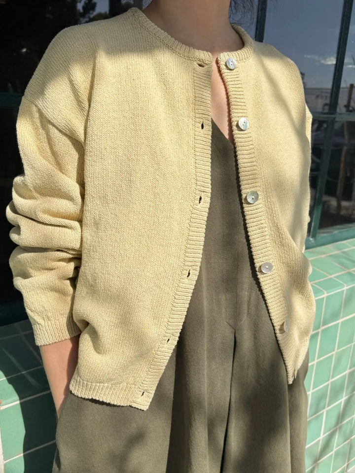 Person wearing a beige 'Cotton Leila Cardigan' in 'Custard' from Le Bon Shoppe over an olive green dress, standing against a tiled wall | Twentyseven Toronto
