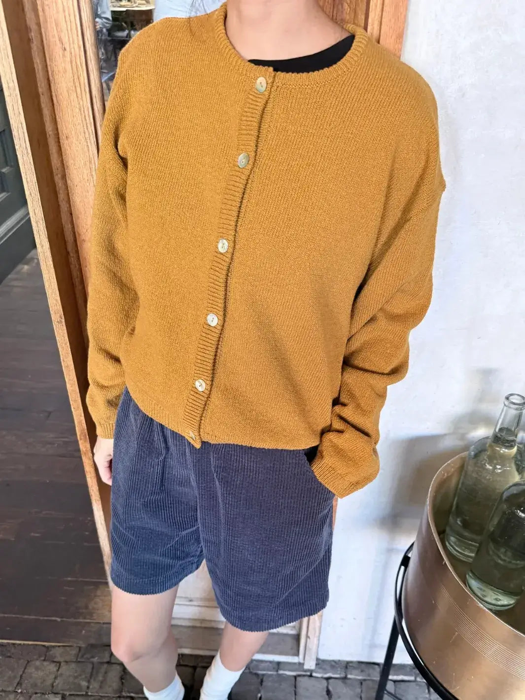 Person wearing a mustard yellow 'Cotton Leila Cardigan' in 'Dijon' with navy shorts from Le Bon Shoppe in an indoor setting | Twentyseven Toronto