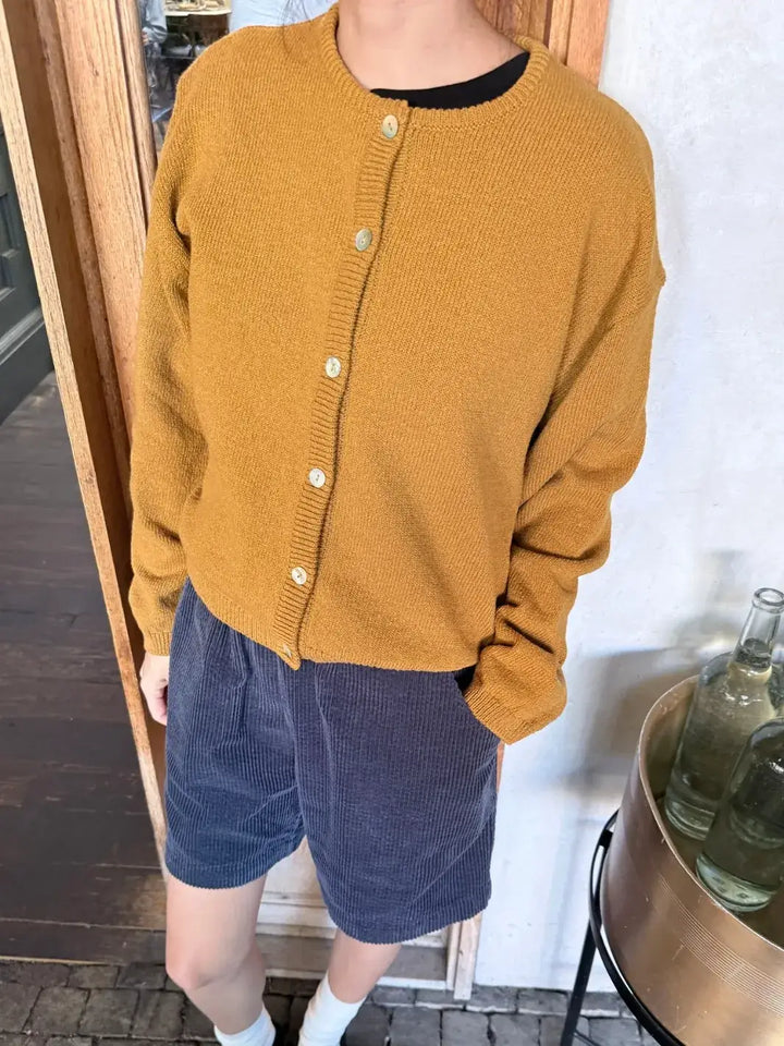 Person wearing a mustard yellow 'Cotton Leila Cardigan' in 'Dijon' with navy shorts from Le Bon Shoppe in an indoor setting | Twentyseven Toronto