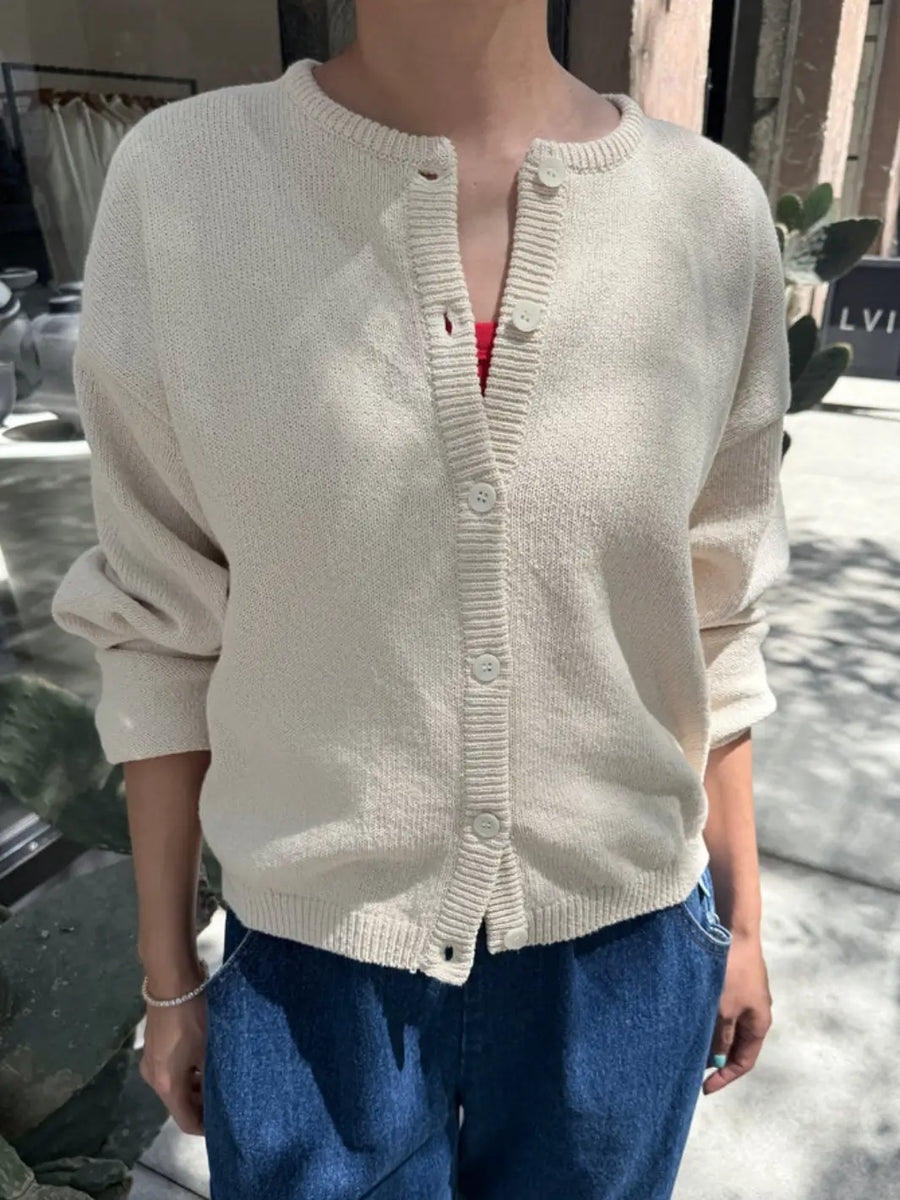 A person wears the cream-colored Le Bon Shoppe Cotton Leila Cardigan - Naturel partially buttoned and paired with relaxed blue jeans | Twentyseven Toronto