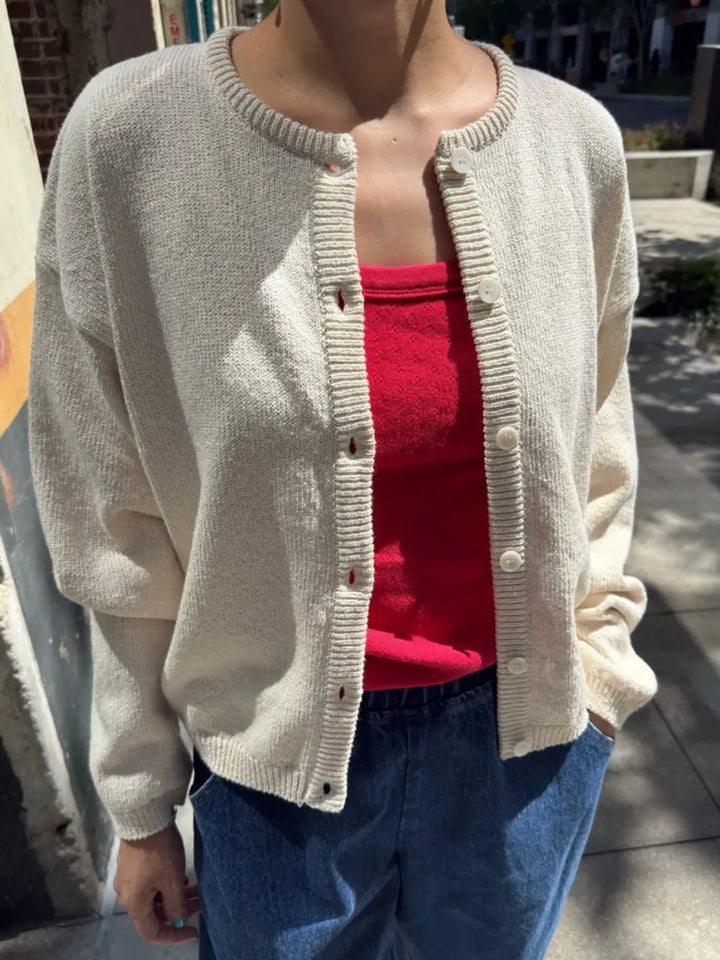 A close-up shot shows a person wearing the unbuttoned Le Bon Shoppe Cotton Leila Cardigan - Naturel layered over a vibrant red top and blue jeans | Twentyseven Toronto