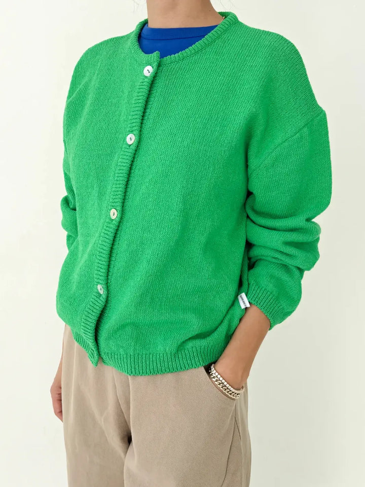 Person wearing a green 'Cotton Leila Cardigan' in 'Spring Green' over a blue shirt with beige pants from Le Bon Shoppe on a white background | Twentyseven Toronto