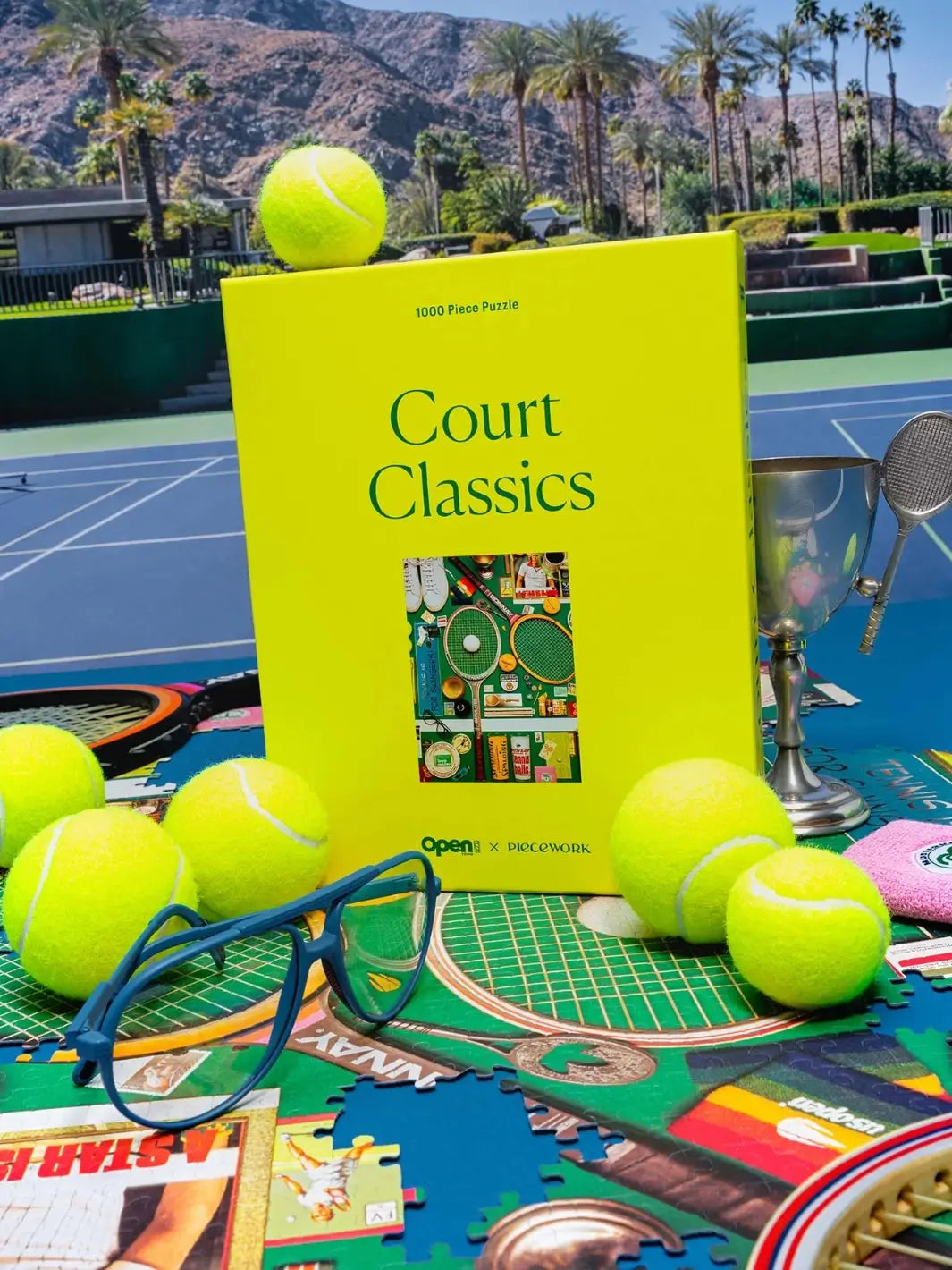 The bright chartreuse box of the Court Classics 1000 Piece Puzzle from Piecework Puzzles sits on an outdoor tennis court surrounded by scattered jigsaw pieces, neon tennis balls, a silver trophy, and blue glasses | Twentyseven Toronto