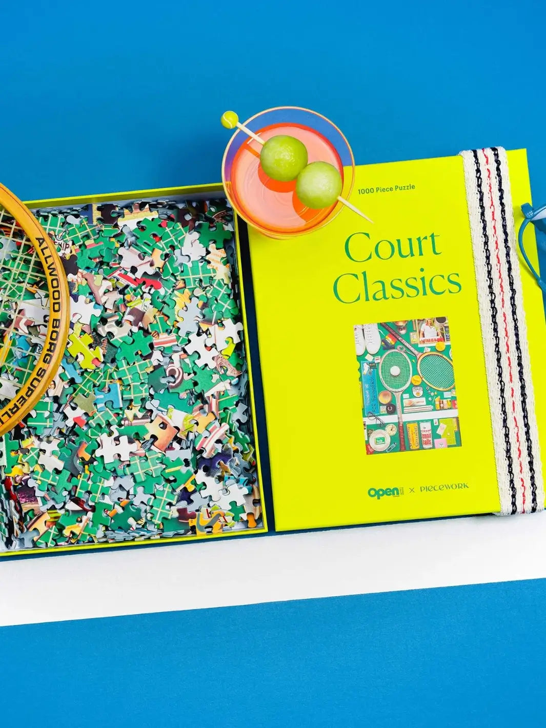 An open box filled with colorful jigsaw pieces next to its chartreuse lid, a tennis racket, and a cocktail garnished with melon balls, highlighting the Court Classics 1000 Piece Puzzle from Piecework Puzzles | Twentyseven Toronto