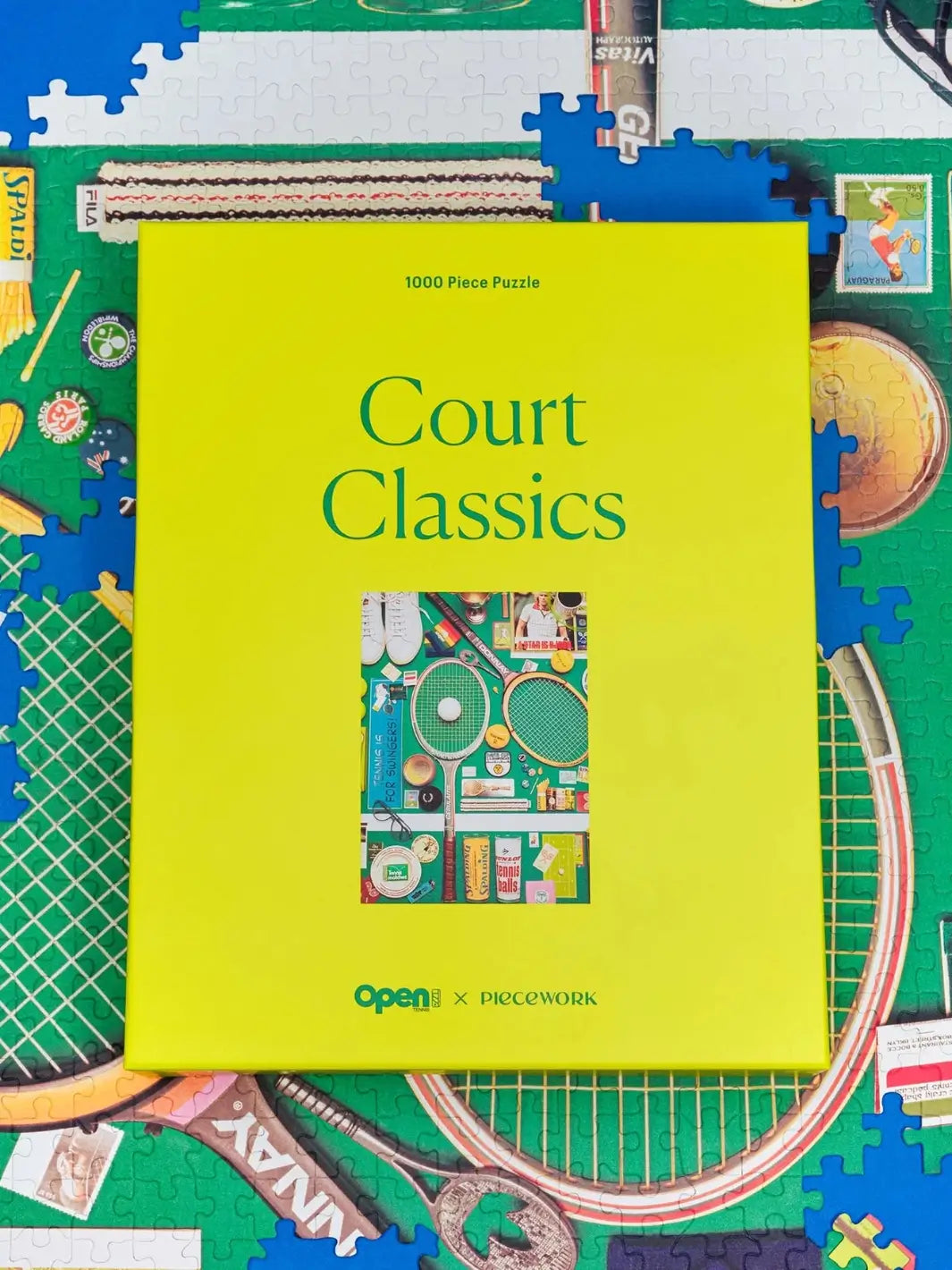 Bright yellow box for the Court Classics 1000 Piece Puzzle from Piecework Puzzles is centered directly on top of its partially completed, matching tennis-themed jigsaw puzzle layout | Twentyseven Toronto