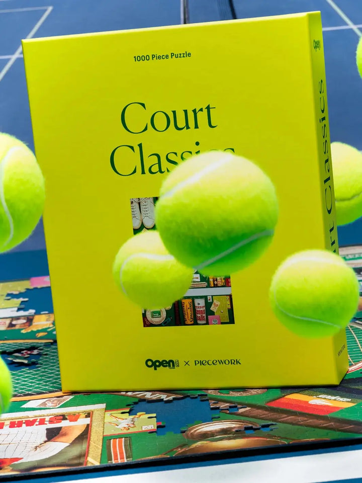 Multiple neon yellow tennis balls bouncing in front oof the chartreuse box for the Court Classics 1000 Piece Puzzle from Piecework Puzzles, which rests on an unfinished puzzle layout | Twentyseven Toronto