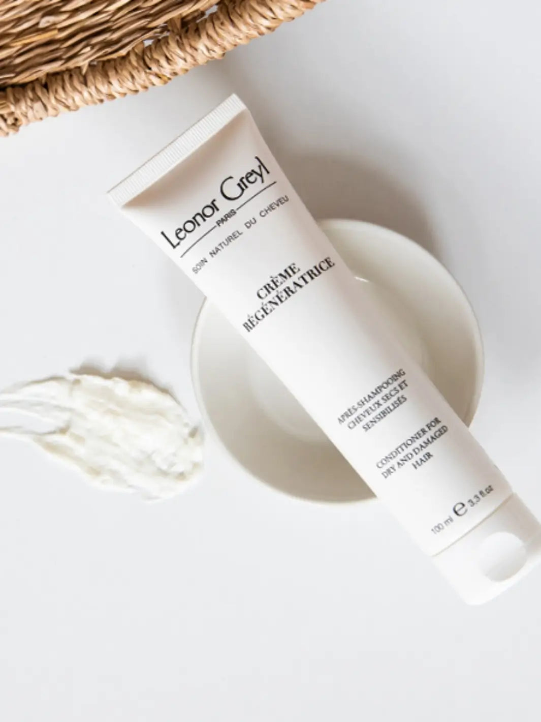 Leonor Greyl 'Crème Régénératrice' conditioner tube on a white surface with a product swatch and wicker basket in the background | Twentyseven Toronto