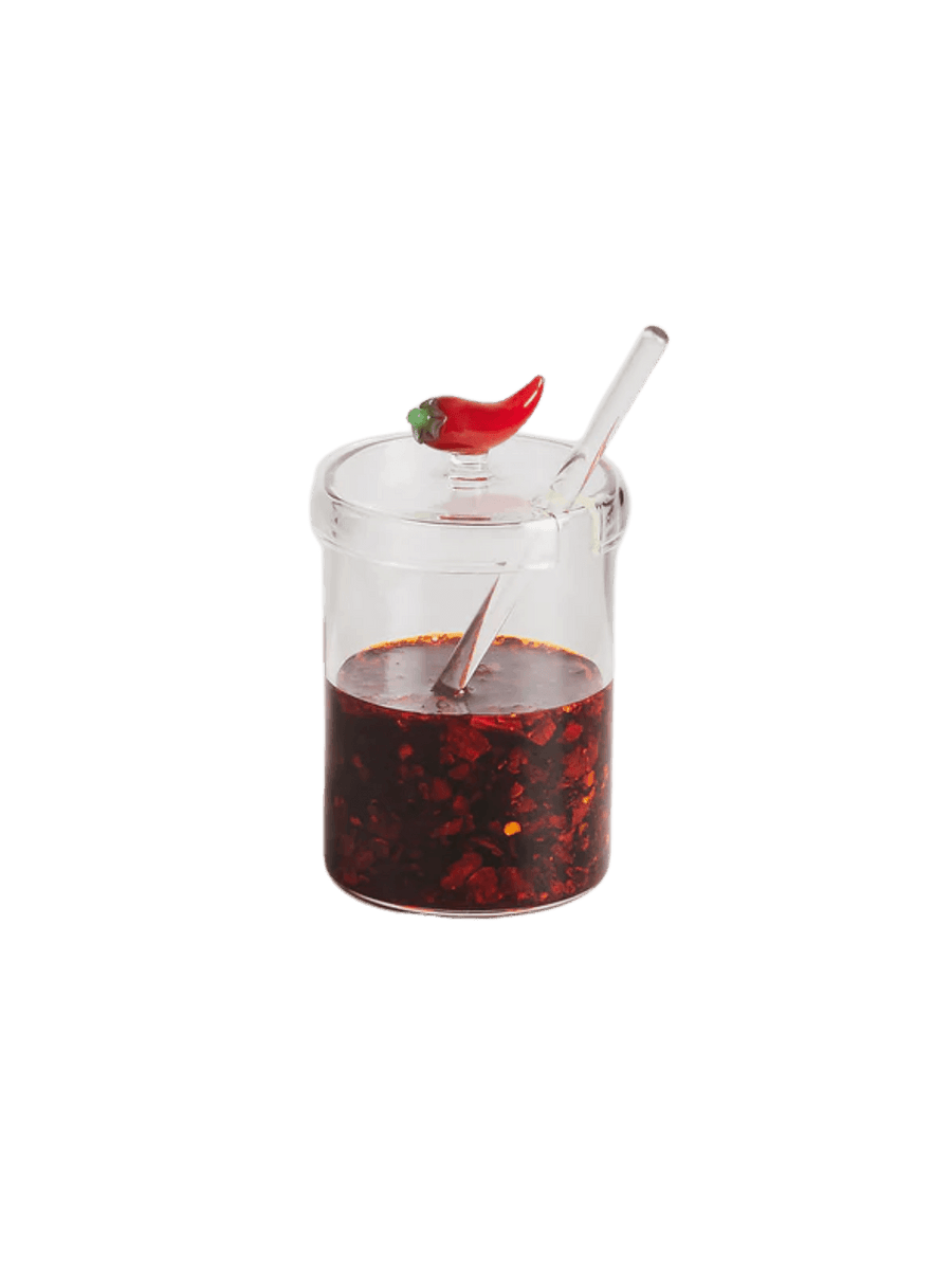 Clear crispy chili oil jar from Maison Balzac with red chili oil inside, a spoon, and a decorative red pepper design on a transparent background | Twentyseven Toronto