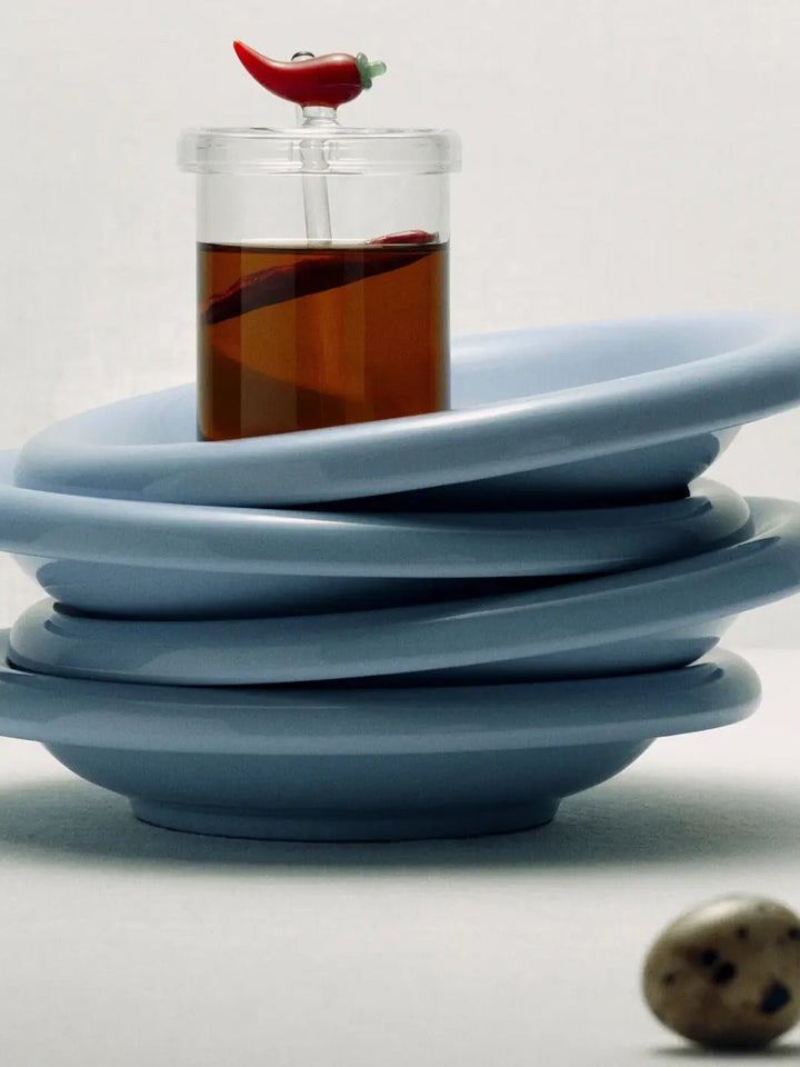 Stack of blue ceramic plates with clear crispy chili oil jar from Maison Balzac with red chili oil inside on top | Twentyseven Toronto