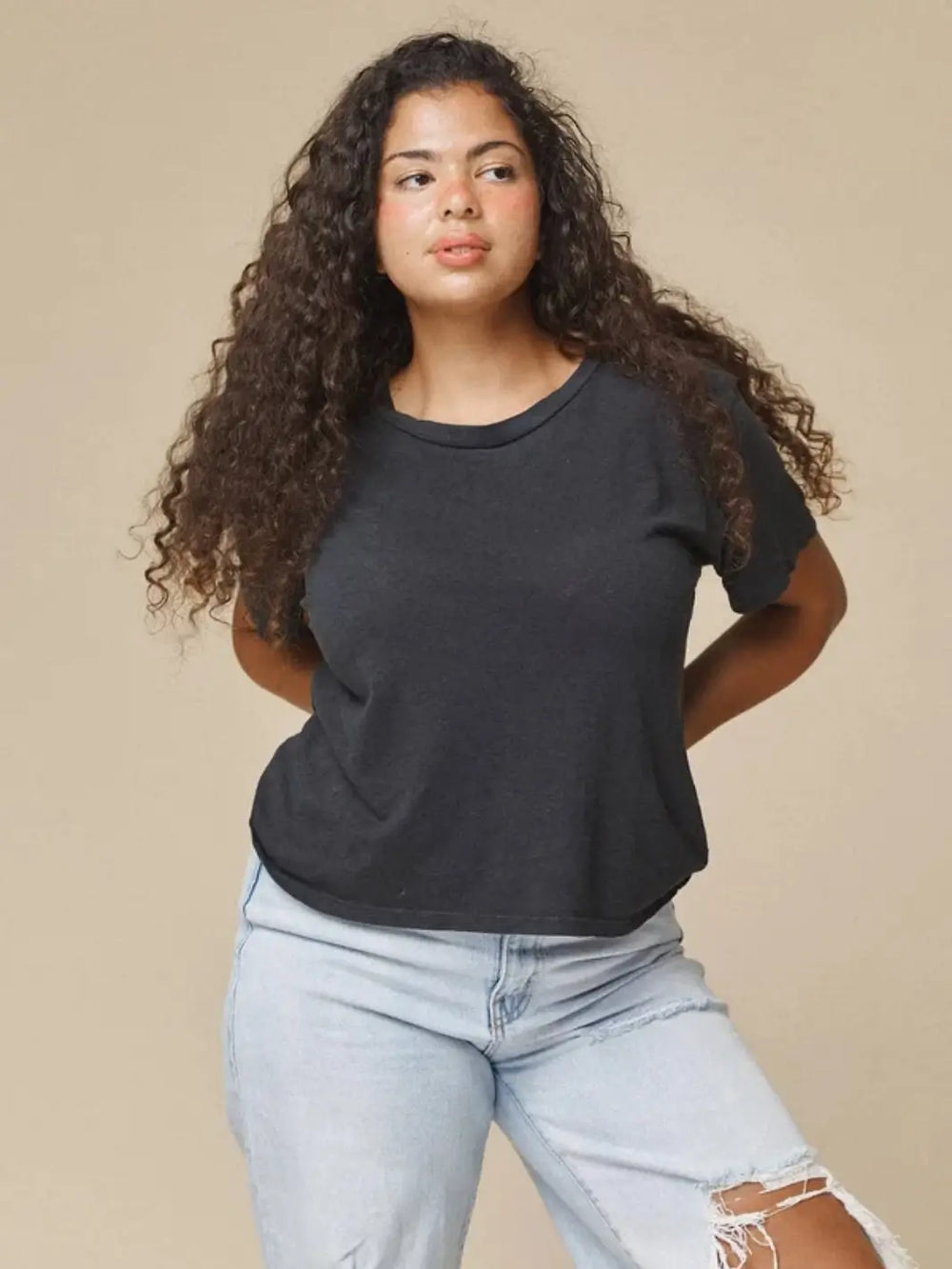 A model with long curly hair wearing the Jungmaven Cropped Lorel Tee in Black paired with light-wash distressed jeans against a beige backdrop | Twentyseven