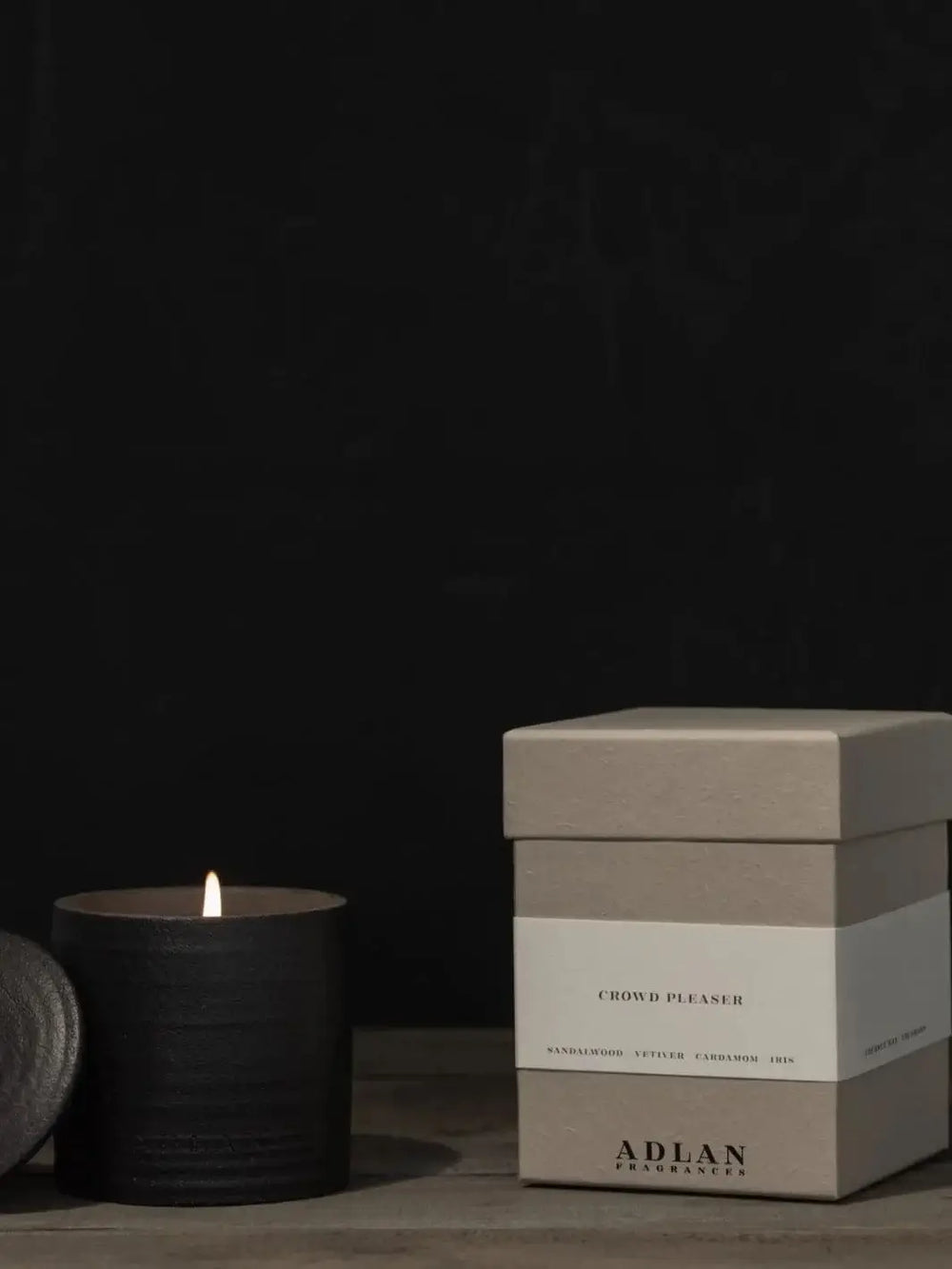 A lit Adlan Fragrances Crowd Pleaser - Petite candle glows inside its textured black jar alongside its minimalist grey product box | Twentyseven Toronto