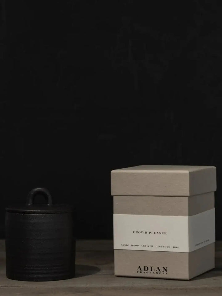 The Adlan Fragrances Crowd Pleaser - Petite candle sits on a wooden surface with its black lid on, positioned next to its square grey packaging box against a dark background | Twentyseven Toronto
