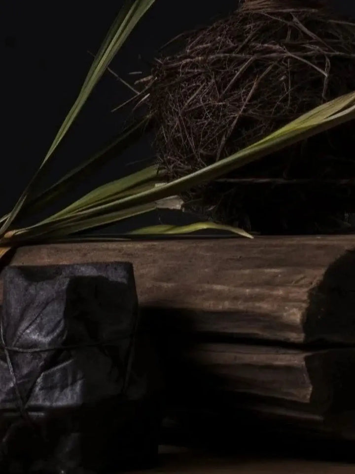 An atmospheric close-up shows rustic wooden blocks, a dark wrapped bundle, and a ball of dried roots with green leaves, representing the natural fragrance notes of the Adlan Fragrances Crowd Pleaser - Petite candle | Twentyseven Toronto