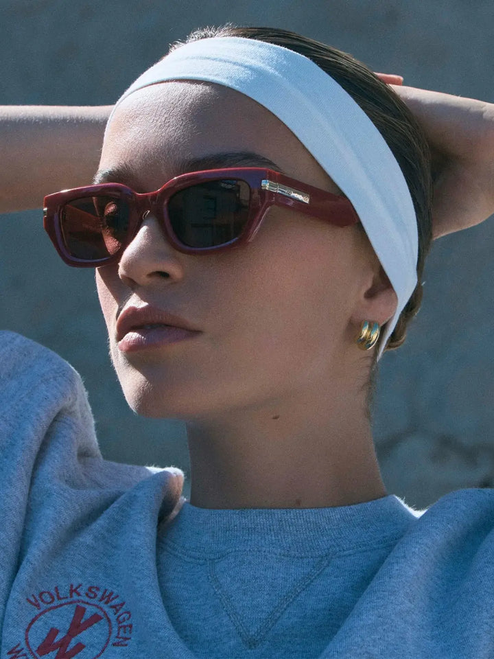 A woman wearing a white headband and thick, rectangular Le Specs Cubito + Burgundy sunglasses in an outdoor setting | Twentyseven Toronto