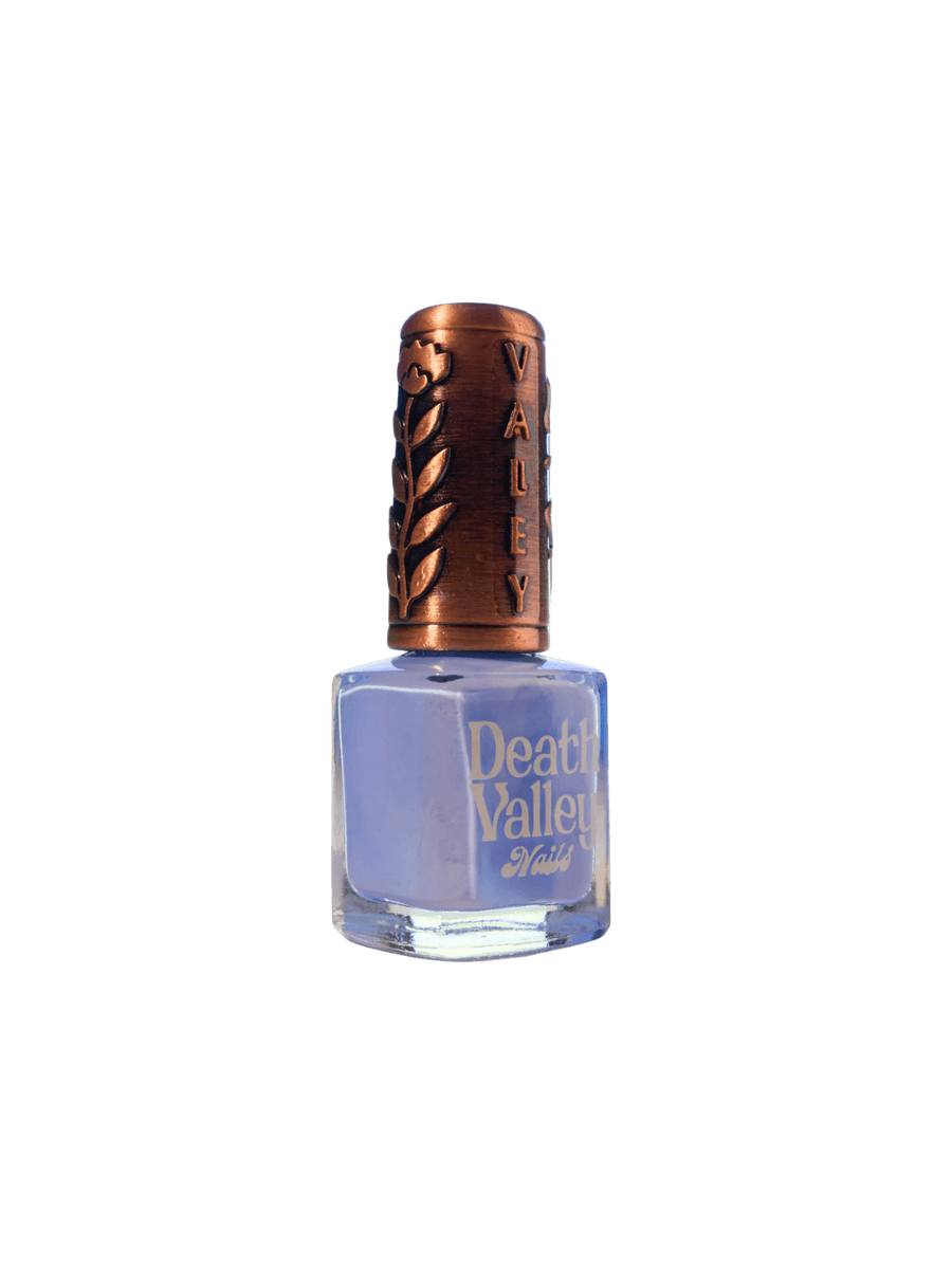 A bottle of Death Valley Nails Curiosity Cloud Nail Polish filled with light cornflower blue lacquer features a decorative copper-toned cap with an embossed floral and brand design | Twentyseven Toronto