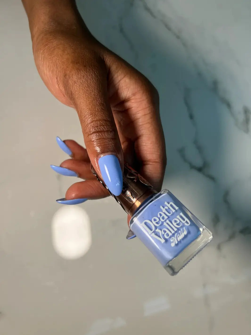 A hand with a deep skin tone holds a bottle of Death Valley Nails Curiosity Cloud Nail Polish, showcasing long, almond-shaped nails painted in a matching soft blue hue | Twentyseven Toronto