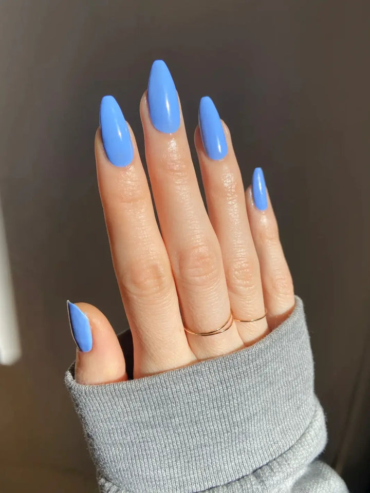 A hand with long, pointed nails painted in light blue Death Valley Nails Curiosity Cloud Nail Polish is shown wearing two thin gold rings against a dark background | Twentyseven Toronto