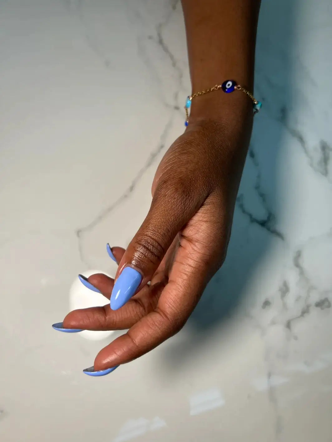 A hand with a deep skin tone and long, stiletto-shaped nails painted in Death Valley Nails Curiosity Cloud Nail Polish rests over a white marble surface | Twentyseven Toronto