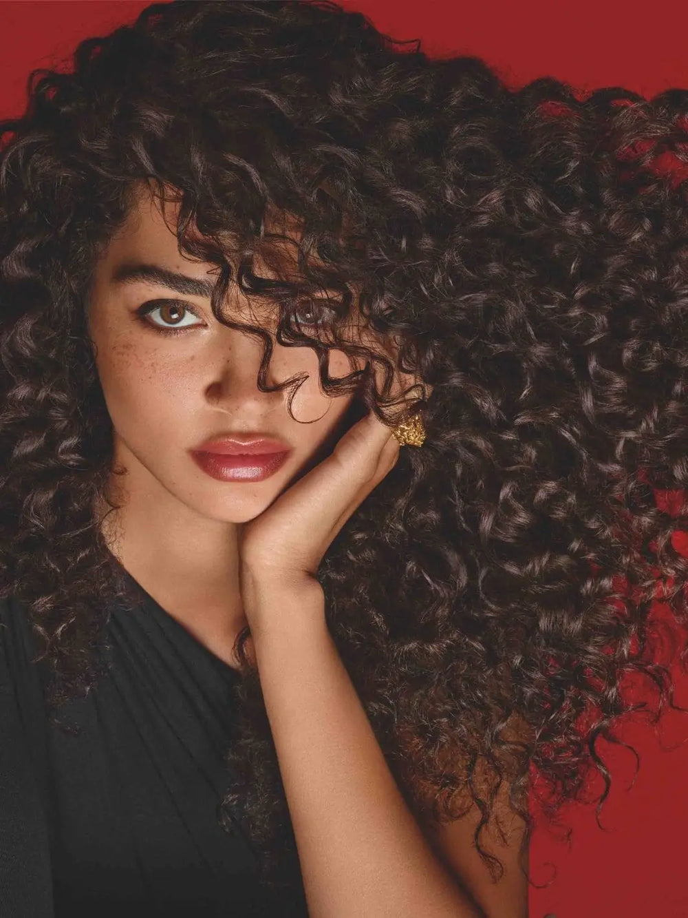 A close-up portrait of a woman with highly defined, voluminous dark curls against a rich red background highlights the hydrating results of Hair by Sam McKnight Curl Cleanse Cleansing Conditioner | Twentyseven Toronto