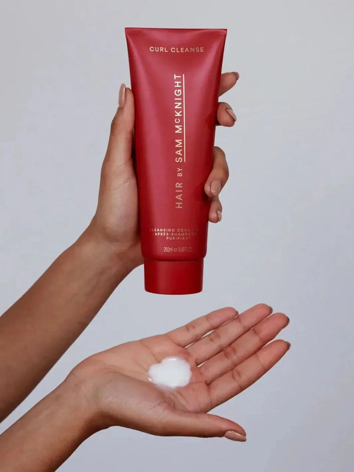 A hand holds a red squeeze tube to dispense a dollop of creamy white product into an open palm, showcasing the texture of Hair by Sam McKnight Curl Cleanse Cleansing Conditioner | Twentyseven Toronto
