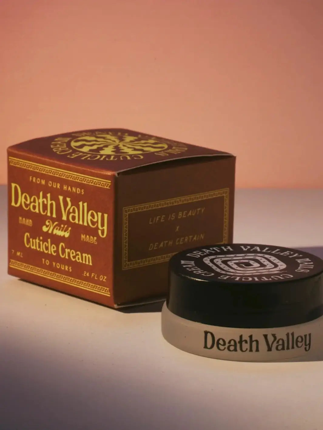Product packaging for Death Valley Nails Cuticle Cream - Eucalyptus + Peppermint + Lavender, consisting of a brown box and a small jar with a black lid, sits on a surface against a soft, warm backdrop | Twentyseven Toronto