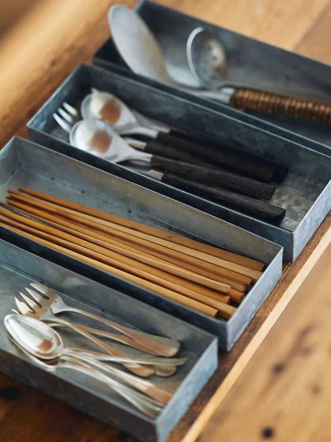 A row of galvanized metal Fog Linen Work Cutlery Box containers neatly organizes wooden chopsticks, spoons, and forks on a rustic wooden table | Twentyseven Toronto