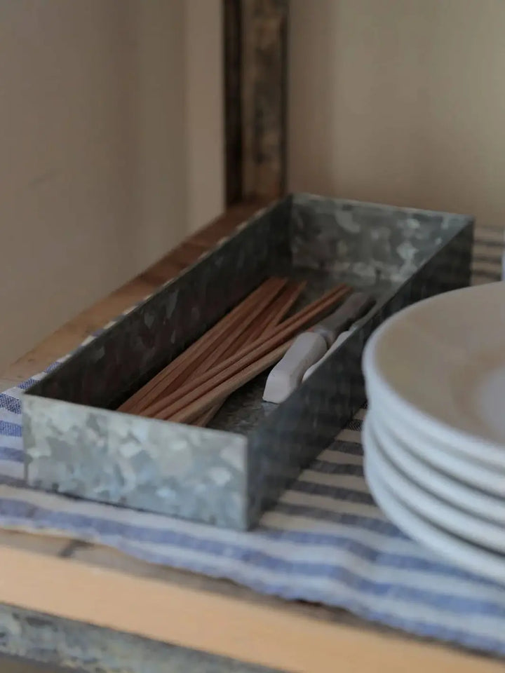 A single Fog Linen Work Cutlery Box containing wooden chopsticks sits on a blue-and-white striped cloth next to a stack of white ceramic plates | Twentyseven Toronto
