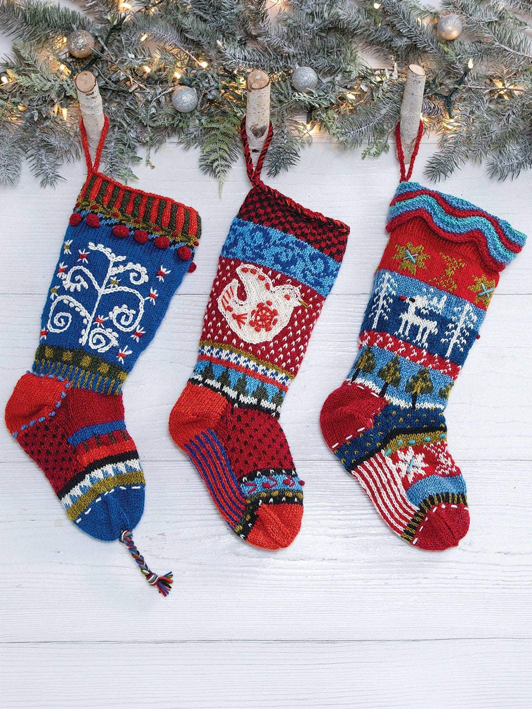 Three colourful Lost Horizons Canada hand-knit wool Christmas stockings hanging beneath festive greenery, featuring Nordic-inspired patterns, reindeer and snowflakes | Twentyseven Toronto