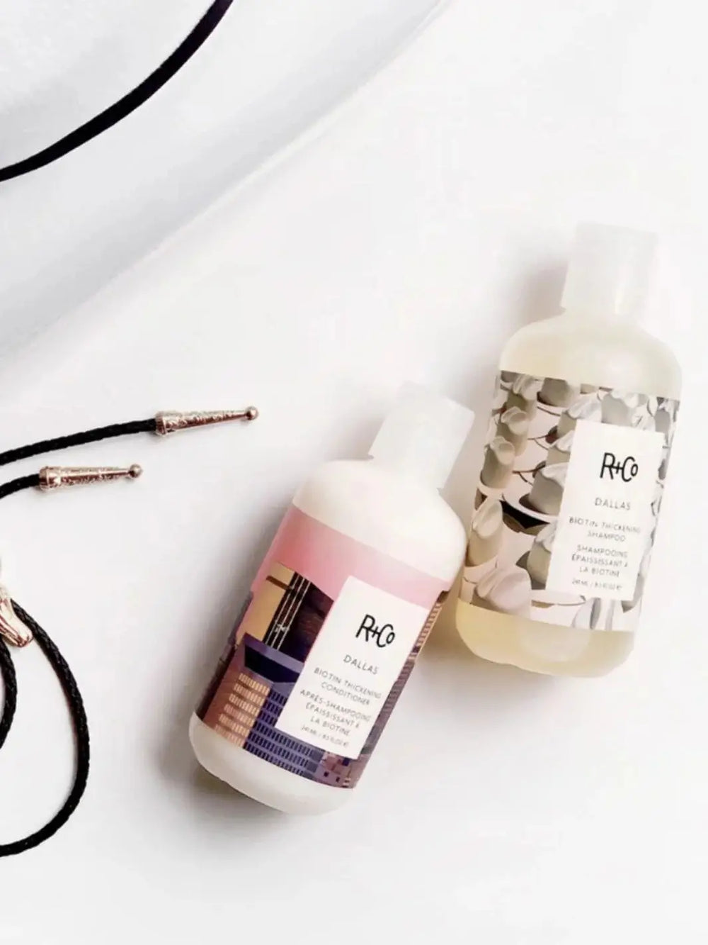 A bottle of R+Co DALLAS Biotin Thickening Shampoo lays flat on a white surface next to its matching conditioner and a black string cord | Twentyseven Toronto