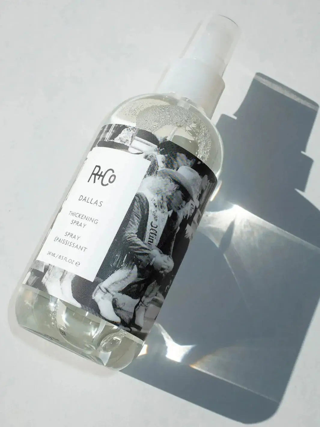A clear spray bottle of R+Co DALLAS Thickening Spray lies flat on a bright surface, casting a crisp shadow | Twentyseven Toronto