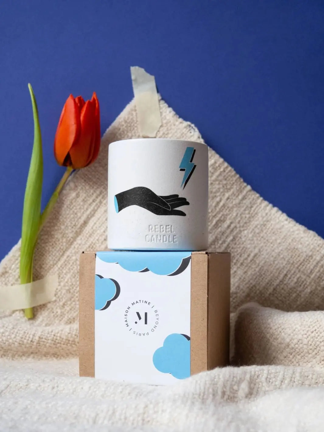 Maison Matine Dark and Stormy Scented Candle with decorative hand, lightning bolt, and cloud packaging and a tulip on a textured surface with a blue background 160g | Twentyseven Toronto