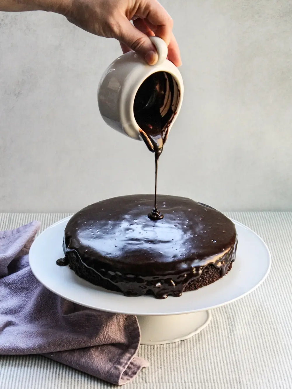 A hand pours glossy chocolate espresso glaze from a small white pitcher onto a freshly baked cake made with the Glazed Cake Co Dark Chocolate Cake Mix | Twentyseven Toronto