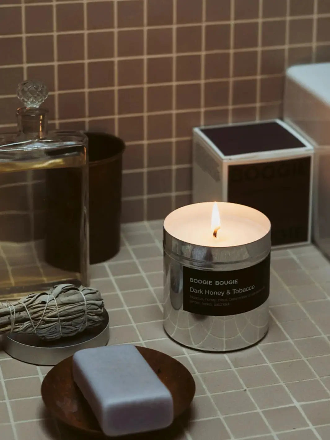 Dark Honey and Tobacco candle in a metallic container with a black 'Boogie Bougie' label on a tiled surface, surrounded by soap and a bundle of sage | Twentyseven Toronto