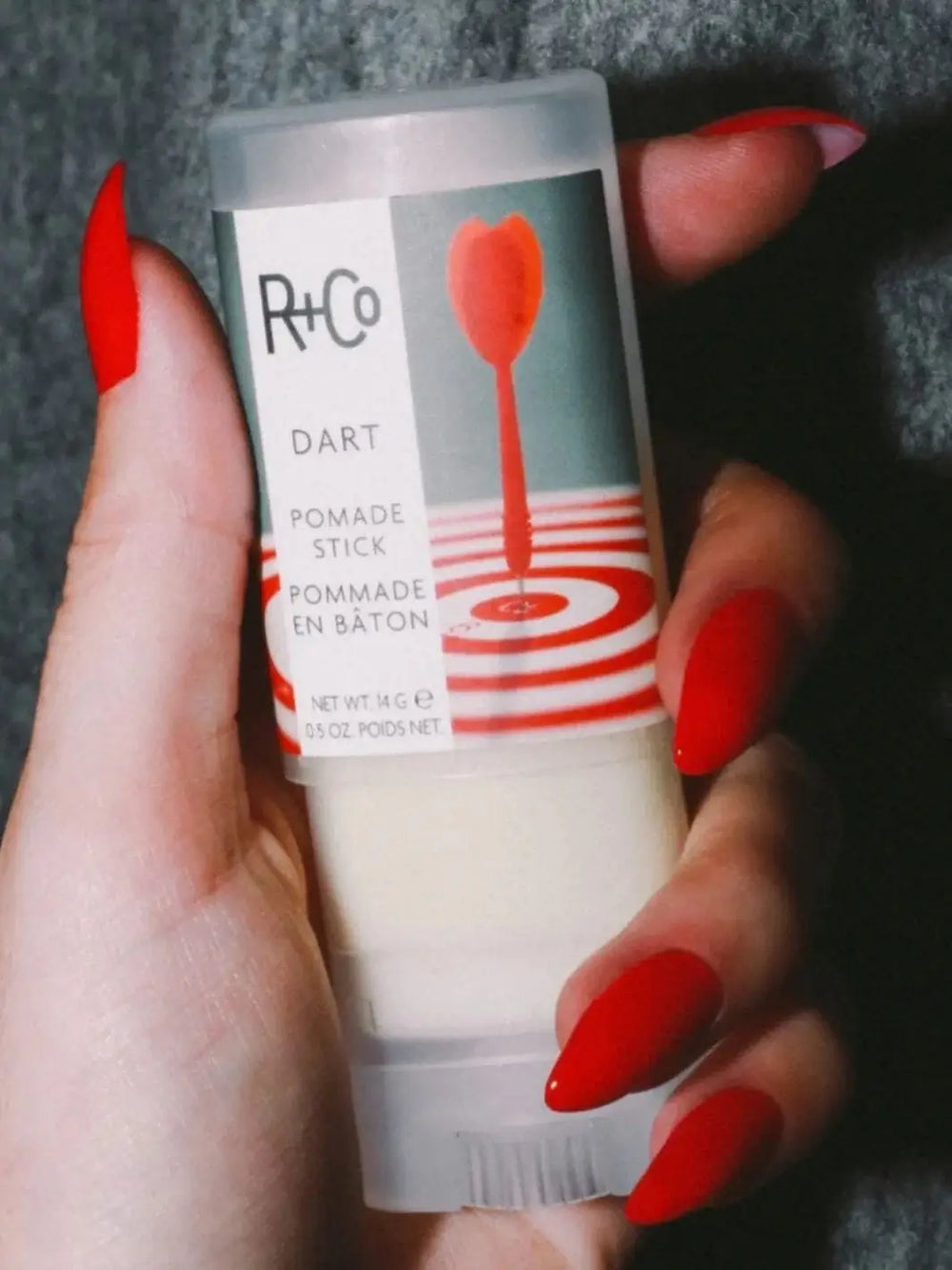 A close-up view of a hand with long, bright red nails holding a container of R+Co DART Pomade Stick against a textured grey background | Twentyseven Toronto