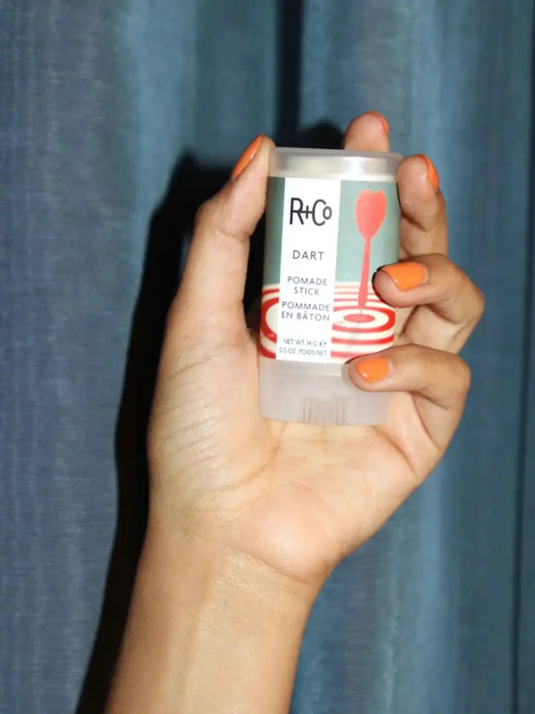 A hand with bright orange nail polish holds up a container of R+Co DART Pomade Stick in front of a blue fabric curtain background | Twentyseven Toronto