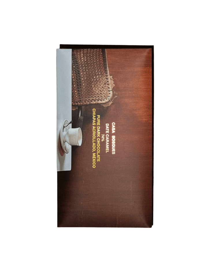 Wrapper of Date Caramel 74% Chocolate Bar from Casa Bosques featuring gold typography over an warm-toned image of a woven chair and a white espresso cup | Twentyseven Toronto