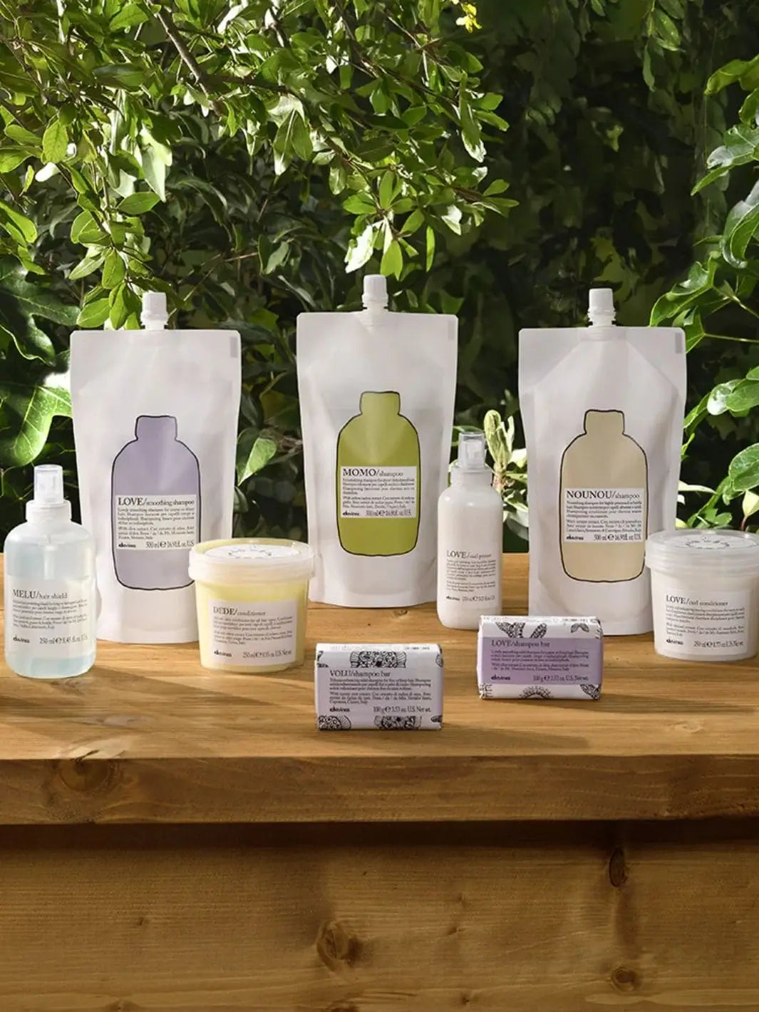Three varieties of Davines Shampoo Refill pouches are arranged on a wooden surface alongside several matching hair care products, tubs, and shampoo bars against a background of green leaves | Twentyseven Toronto