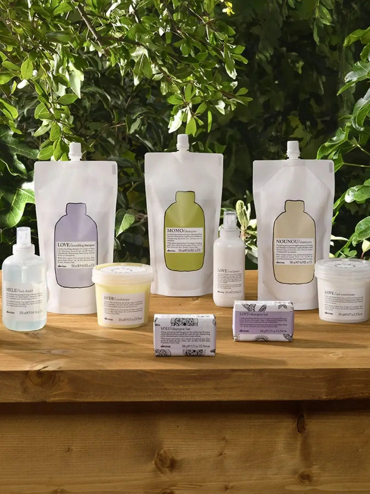 Three varieties of Davines Shampoo Refill pouches are arranged on a wooden surface alongside several matching hair care products, tubs, and shampoo bars against a background of green leaves | Twentyseven Toronto