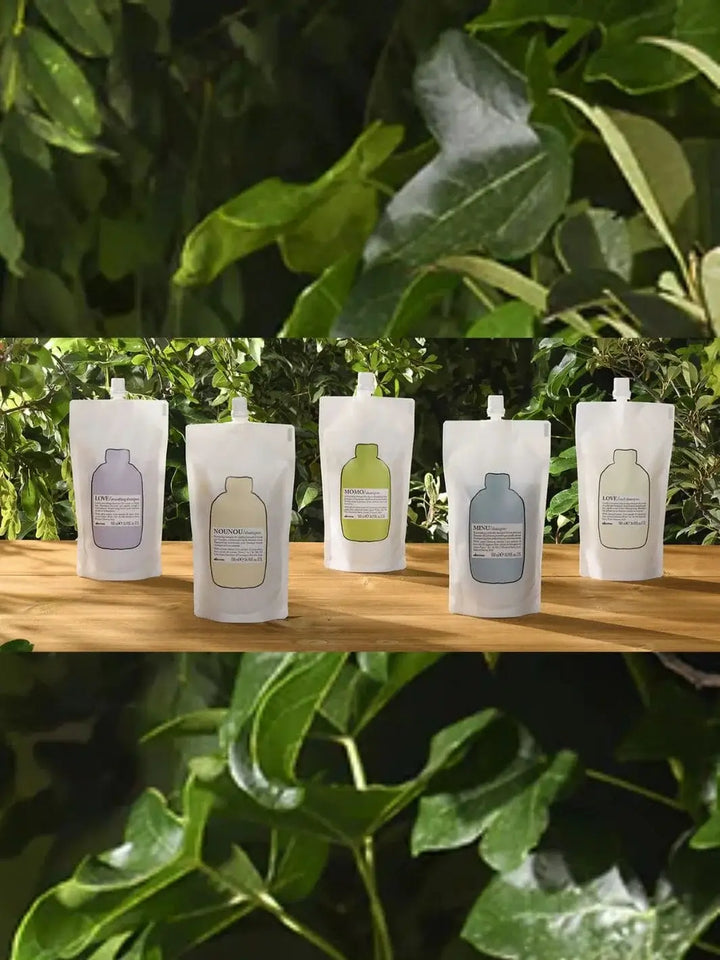 Five different varieties of Davines Shampoo Refill pouches stand lined up on a sunny wooden platform, framed by green leaves on the top and bottom | Twentyseven Toronto