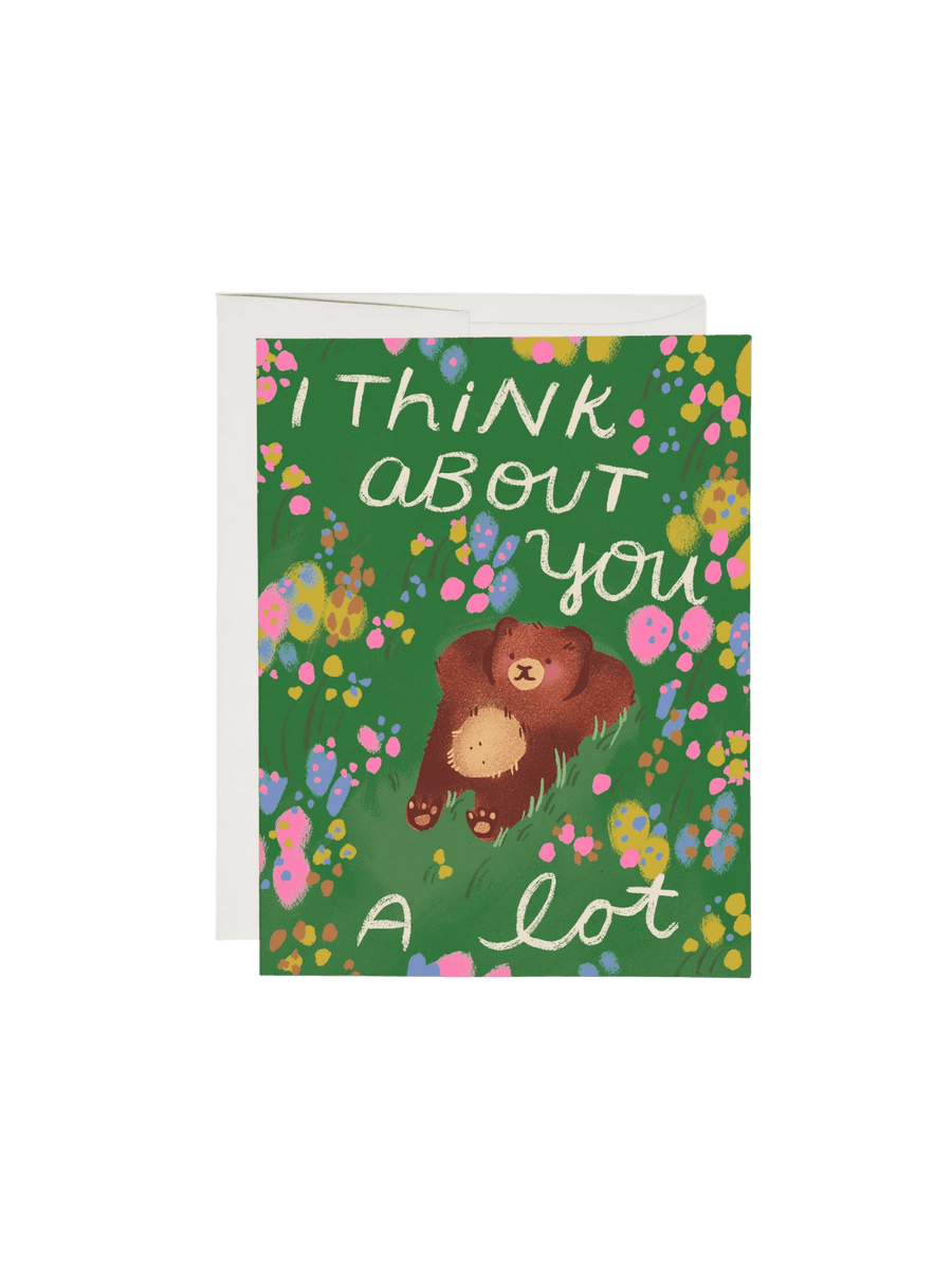 A Daydreaming Greeting Card by Dylan Mierzwinski for Red Cap Cards features an illustration of a brown bear lying on his back in a grassy field of flowers beneath the text "I think about you a lot," next to a white envelope | Twentyseven Toronto